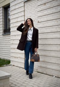 Femme debout près d'un mur en briques blanches, portant un manteau marron foncé, un chemisier blanc, un jean bleu, des bottes noires, tenant un sac à main marron.