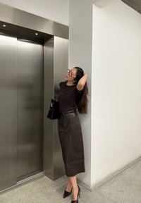 Brun læder midi-nederdel, sort tætsiddende top, transparente briller og høje hæle, står ved siden af en metal elevator i et moderne rum.