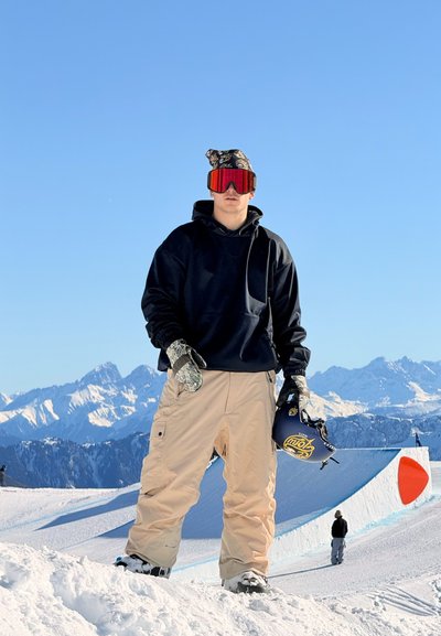 Un snowboarder con una sudadera negra y pantalones de nieve color arena está de pie sobre la nieve, llevando gafas rojas con espejo y guantes con estampados, sosteniendo un casco azul.