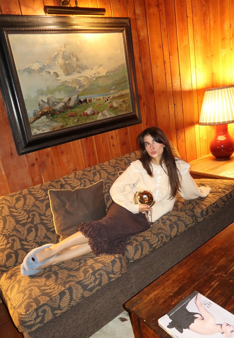 Vilnonė sijonas su nėrinių apdaila, smėlio spalvos megztinis su nėrinių akcentais, puošnus sofa su lapų raštu, medinis stalas, kalnų paveikslas fone.