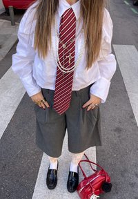 Camisa branca cropped, gravata riscada em borgonha, calções cinzentos plissados, mocassins pretos, meias brancas, colares de pérolas em camadas, bolsa vermelha com ornamentação de flor.