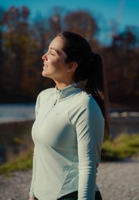 Hosszú copfot viselő nő világoskék cipzáros felsőben, csukott szemmel, mosolyogva, szabadban, víz és fák közelében, napsütésben áll.