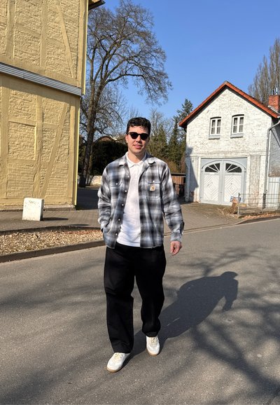 Camisa a cuadros ligera en gris y azul sobre una camiseta blanca, pantalones negros y zapatillas blancas. El fondo presenta edificios y árboles.