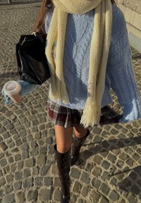 Femme portant un pull en maille torsadée bleu clair, une écharpe jaune, une jupe à carreaux et des bottes noires montantes, tenant une tasse de café et un sac noir dans une rue pavée.