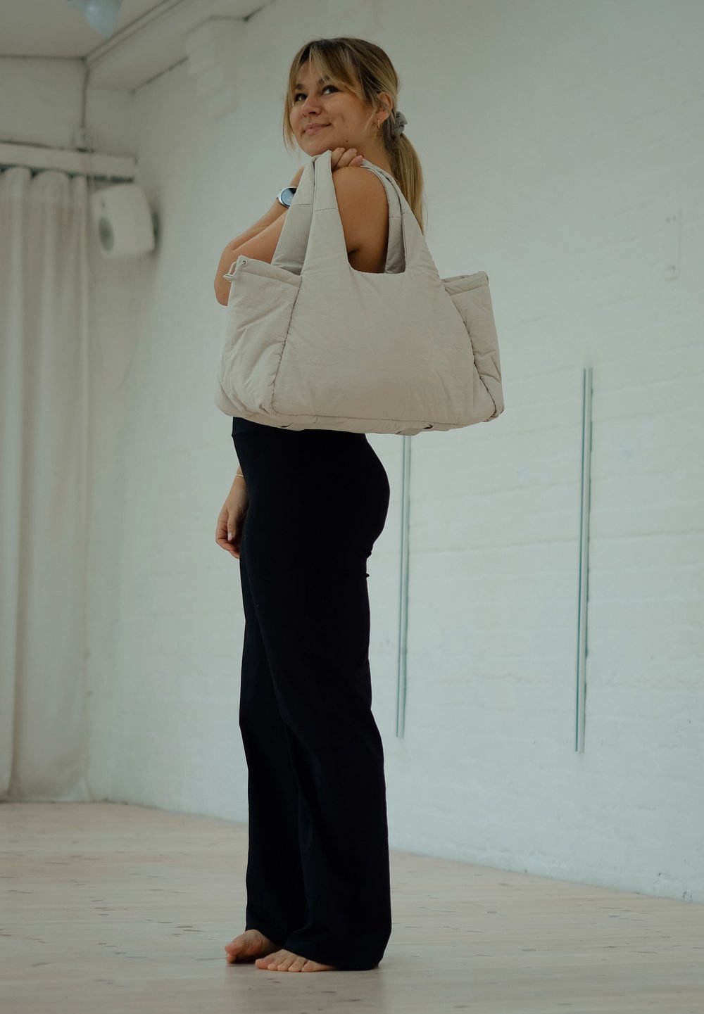 Jeune femme en tenue noire debout pieds nus à l'intérieur, portant un grand sac fourre-tout matelassé beige sur l'épaule et regardant vers le haut.