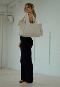 Jeune femme en tenue noire debout pieds nus à l'intérieur, portant un grand sac fourre-tout matelassé beige sur l'épaule et regardant vers le haut.