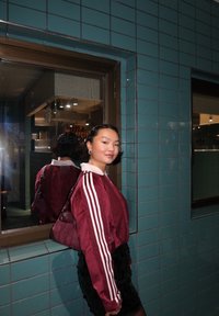 Chaqueta deportiva granate con rayas blancas, combinada con una falda negra texturizada. El fondo presenta azulejos verde azulado y una ventana reflectante.