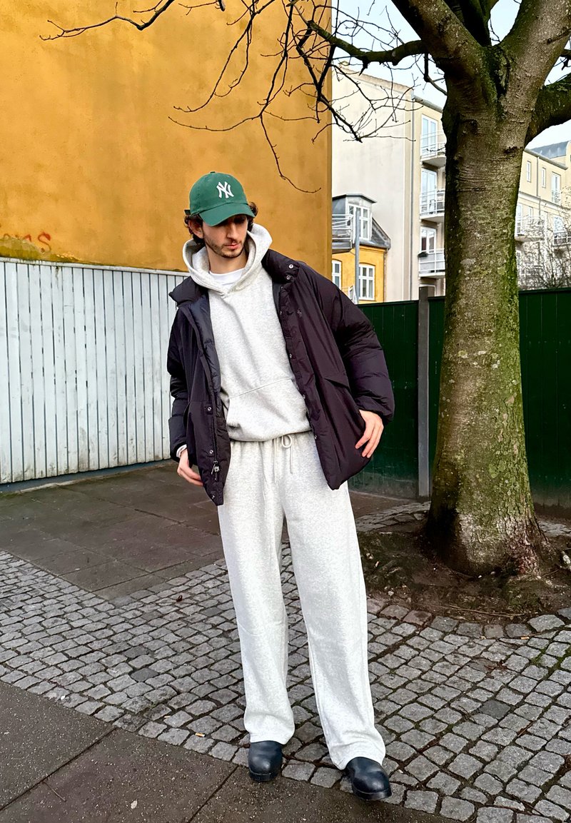 Veste matelassée noire portée sur un sweat à capuche gris et un pantalon de jogging assorti. Casquette NY verte, sabots foncés, et pavés en pierre dans un cadre urbain.