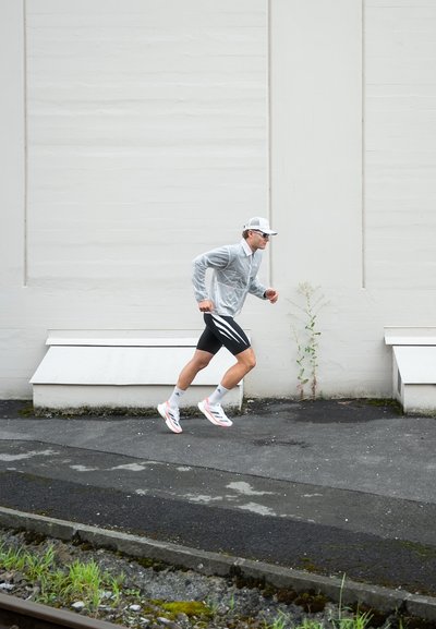 Hombre atlético corriendo con una chaqueta cortavientos gris claro y pantalones cortos negros con rayas blancas. Lleva una gorra blanca y zapatillas de correr coloridas.
