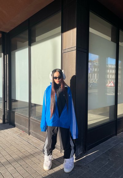 Cazadora azul con rayas blancas, camiseta negra, pantalones deportivos negros con rayas, botas blancas, auriculares blancos, cabello largo y oscuro. Entorno urbano.