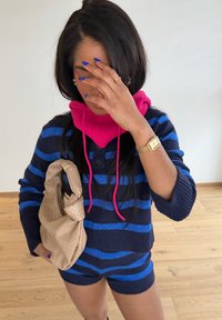 Femme en pull à rayures bleues et shorts, écharpe rose, tenant un sac à main beige, couvrant son visage avec la main montrant des ongles bleus et une montre en or.