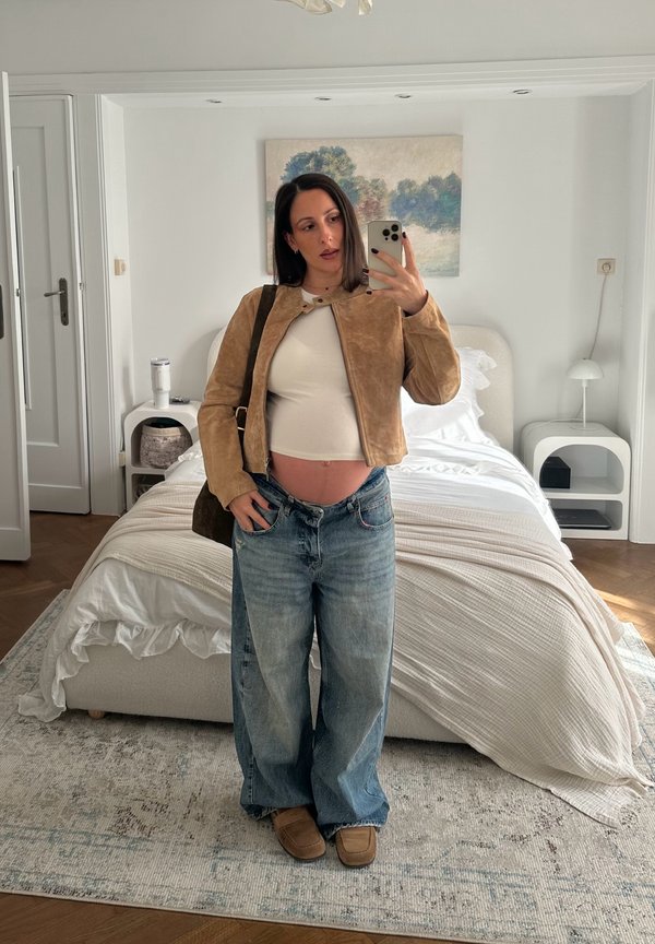 Mujer embarazada con ropa informal tomando una selfie frente al espejo en un dormitorio luminoso con una cama, mesitas de noche y una pintura de paisaje.