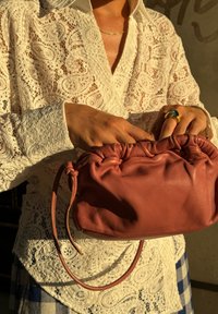 Τσάντα ροζ δέρματος με μαζεμένο επάνω μέρος και ρυθμιζόμενο λουράκι, που ξεκουράζεται χέρι σε χέρι πάνω από μια λευκή δαντελένια μπλούζα και ύφασμα καρό φούστας.