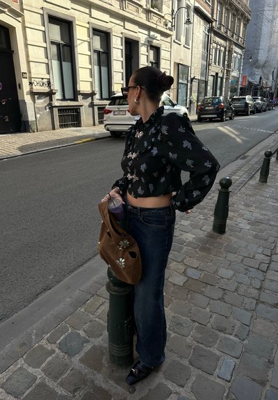 Blusa negra recortada con adornos florales, jeans azules y bolso marrón. Los accesorios visibles incluyen gafas de sol y un vaso de bebida.