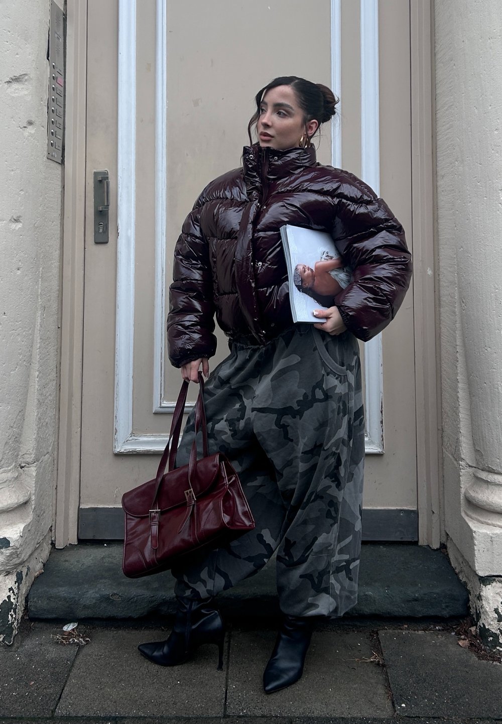 Glossy burgundy padded jacket, grey camouflage skirt, black heeled boots, and a burgundy handbag. Model holding a magazine.