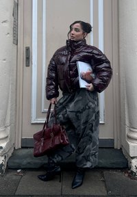 Glossy burgundy padded jacket, grey camouflage skirt, black heeled boots, and a burgundy handbag. Model holding a magazine.