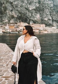Trench coat beige chiaro sopra un top lavorato a maglia bianco e pantaloni larghi neri. Borsa nera in mano, in piedi vicino all'acqua con montagne sullo sfondo.