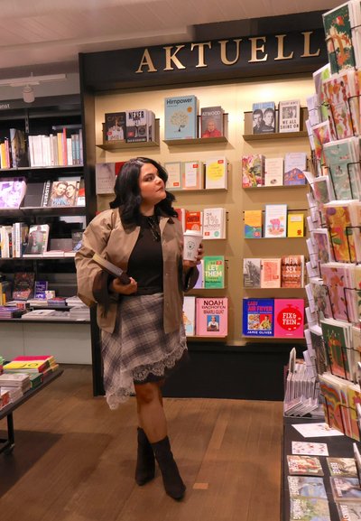 Mujer sosteniendo un libro y una taza de café mientras busca libros en estantes etiquetados como "AKTUELL" en una librería con suelo de madera.
