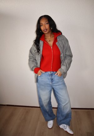 Chaqueta bomber gris sobre suéter rojo de punto con cremallera, combinado con vaqueros de pierna ancha azul claro y zapatillas blancas. Accesorios mínimos.
