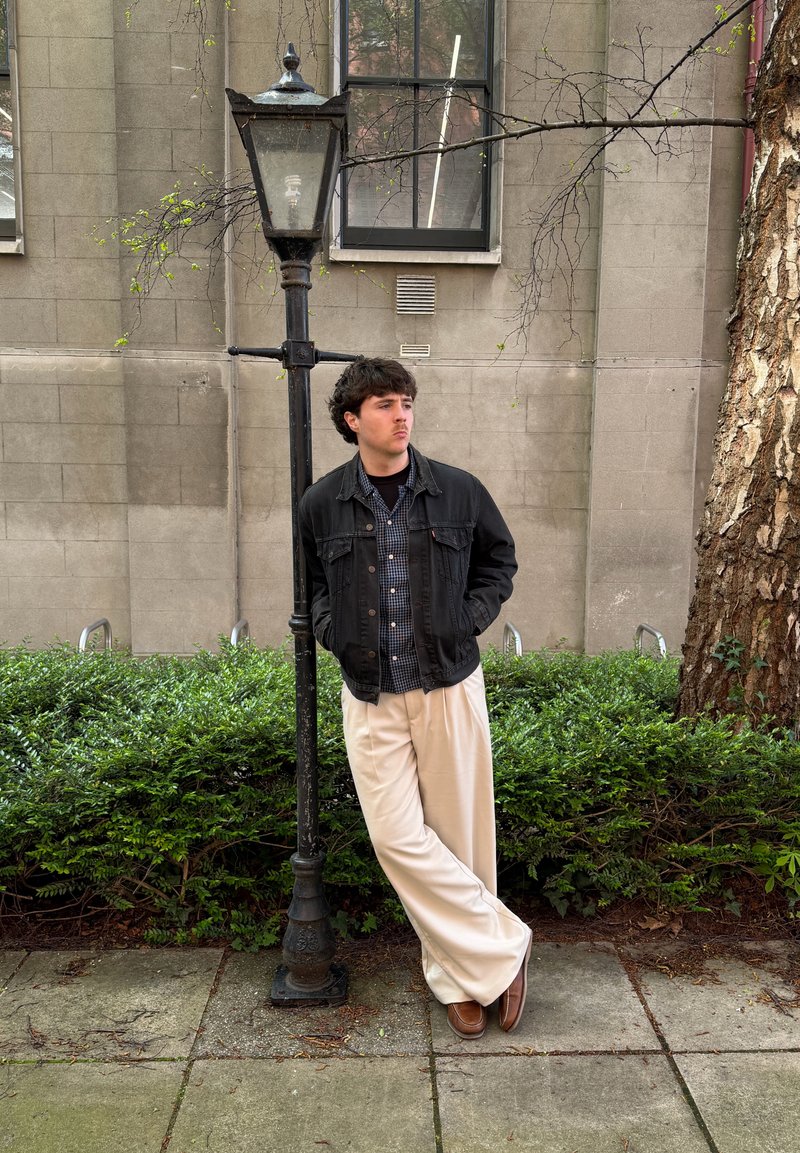 Young man in black jacket and beige pants leaning against a vintage streetlamp beside a building and greenery.