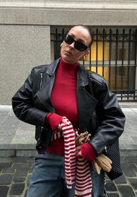 Czarna skórzana kurtka motocyklowa, czerwona bluzka z długim rękawem, szare dżinsy, pasiasty szalik, brązowe rękawiczki oraz czarne okulary przeciwsłoneczne ze złotymi akcentami.