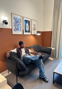 Person med briller og lue som sitter i kors på en mørkegrå sofa i et moderne rom med brune og hvite vegger og innrammet abstrakt kunst.