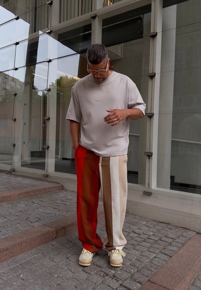 Camiseta de color beige claro, corte relajado; pantalones anchos multicolores con franjas rojas, marrones y crema; zapatillas casuales de color beige; escenario urbano con fondo de cristal.