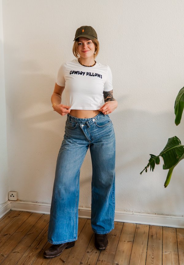 Tânără purtând un tricou alb scurt „Cowboy Pillows”, blugi largi, cizme închise la culoare și o șapcă verde, stând în interior lângă o plantă.