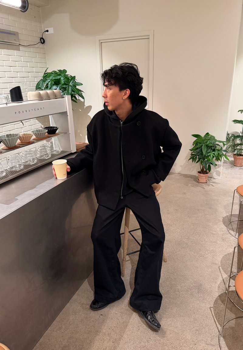 Homme en tenue noire assis sur un tabouret, tenant un gobelet en papier au comptoir d'un café minimaliste avec des verreries et des plantes.