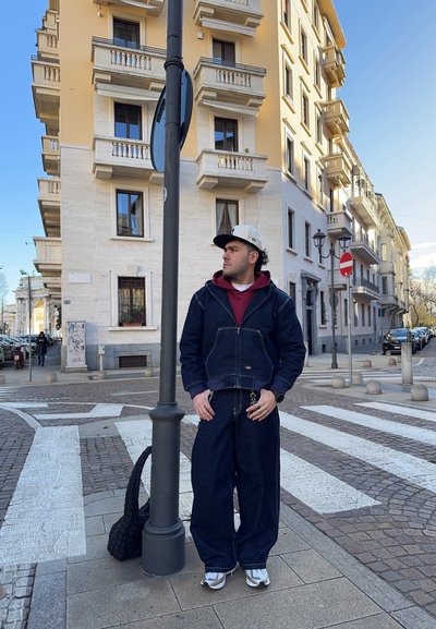 Chaqueta de denim y pantalones, azul oscuro, de corte holgado. Gorra blanca, sudadera roja por debajo, zapatillas plateadas, de pie junto a un poste en un entorno urbano.