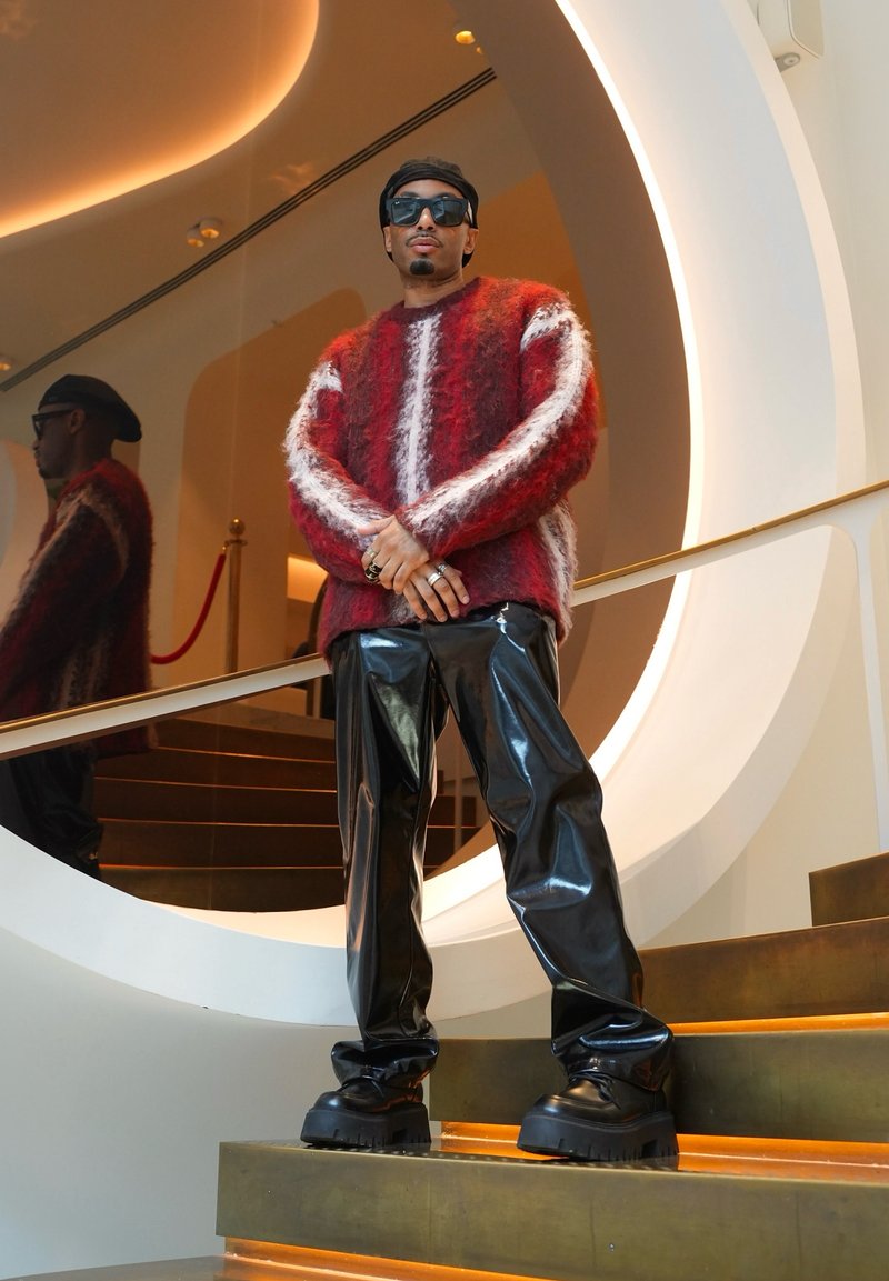 Homme portant de grandes lunettes de soleil noires, un pull pelucheux rouge et blanc, un pantalon noir brillant et des bottes noires épaisses, debout sur des escaliers près d'un miroir circulaire.