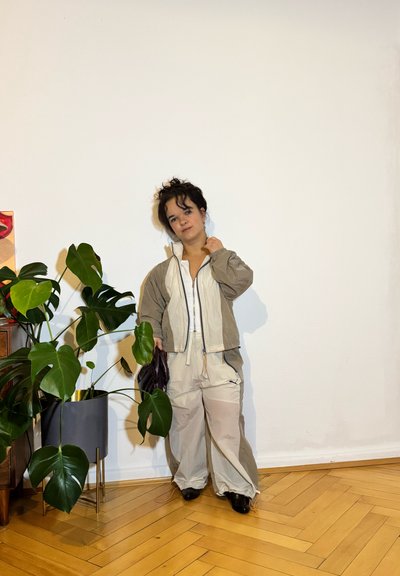 Conjunto en beige claro y verde oliva con pantalones holgados y chaqueta con cremallera. Textura brillosa, con cordón y bolsillos. Zapatos negros. Planta de monstera cerca.