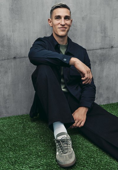 Conjunto informal que incluye una chaqueta marina, un suéter verde oliva, pantalones oscuros y zapatillas grises. Texturas suaves y detalles sutiles en los zapatos.