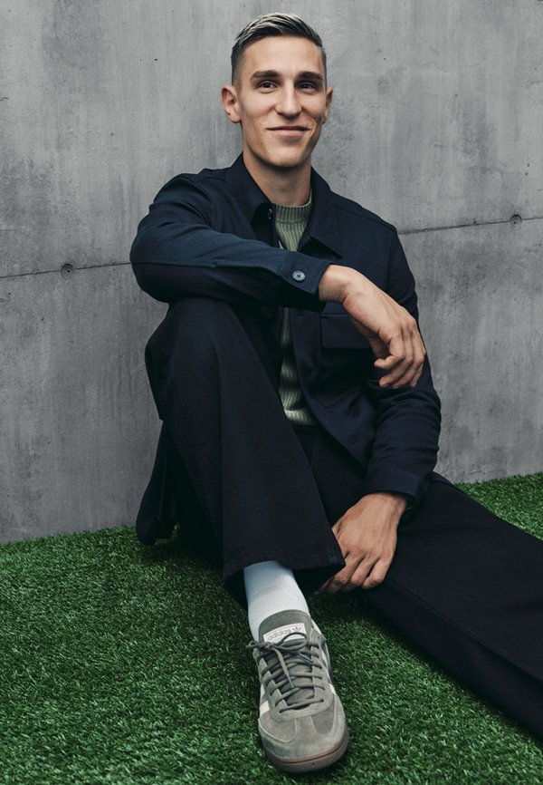 Lässiges Outfit mit einer marineblauen Jacke, einem olivgrünen Pullover, dunklen Hosen und grauen Turnschuhen. Weiche Texturen und dezente Details an den Schuhen.