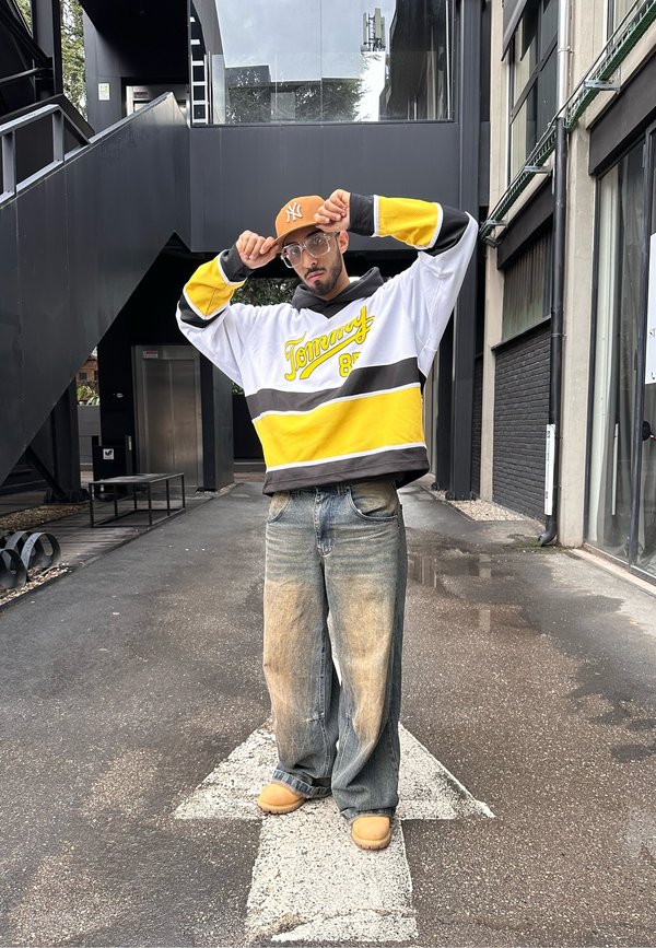 Hoodie oversized bianco, giallo e nero con il testo "Tommy". Indossato con jeans azzurri sbiaditi e zoccoli color sabbia, in piedi su un vialetto pavimentato.