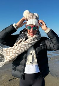 Doudoune noire, pull en tricot beige, bonnet crème avec pompons, écharpe à motif animal, et lunettes de soleil roses reflètent la lumière du soleil.
