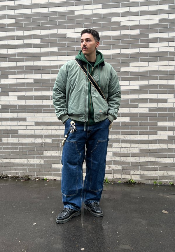 Homme avec moustache portant une veste bomber verte et un sweat à capuche, jean bleu large avec porte-clés, chaussures noires, debout contre un mur de briques grises.