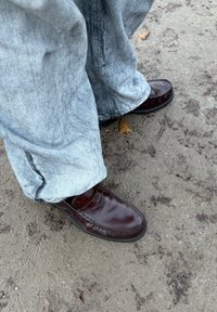 Mocassini marroni in pelle con finitura liscia, abbinati a pantaloni in denim grigio chiaro. Il terreno è terroso e irregolare con foglie visibili.