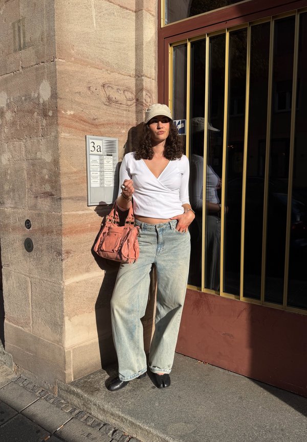 Mujer con top blanco, vaqueros anchos azul claro y gorra beige, apoyada en una pared de piedra cerca de una puerta con barras metálicas, sosteniendo un bolso rosa.
