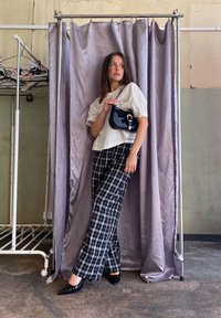 Biely oversize tričko, čierno-biele kárované široké nohavice, čierne špicaté podpätky, drží čiernu kabelku so striebornými akcentmi, pred závesným pozadím.