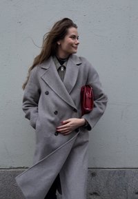 Grigiato cappotto doppiopetto con grandi bottoni, indossato sopra un indumento verde, mentre tiene una piccola borsa rossa. Texture liscia e design fluido.