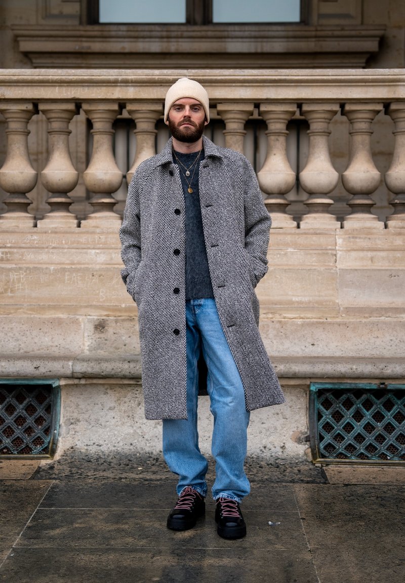 Homme portant un pardessus gris à motifs, un jean bleu clair, des chaussures noires avec des lacets roses, un pull gris et un bonnet beige, debout à l'extérieur.