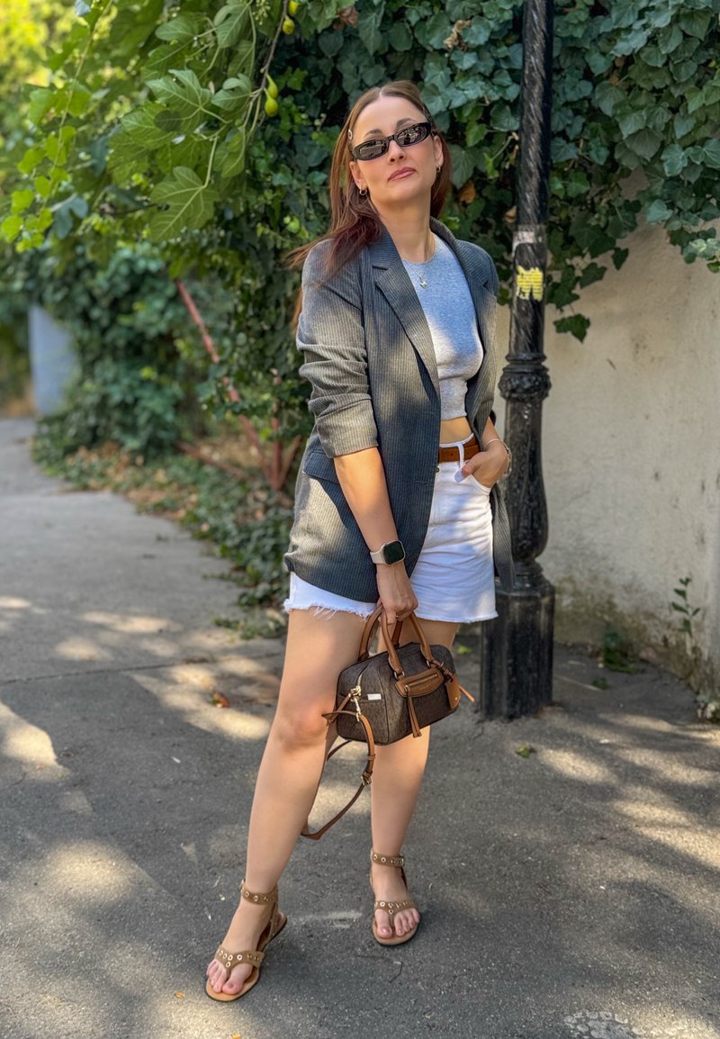 Femme portant des lunettes de soleil, un blazer gris, une jupe blanche et des sandales, se tenant sur le trottoir avec un petit sac à main marron près de la verdure feuillue et d'un lampadaire.