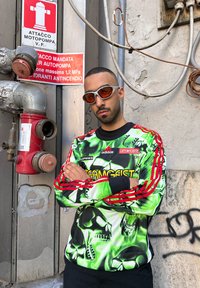 Άνδρας με κοντά μαλλιά και πορτοκαλί γυαλιά ηλίου, που φοράει πράσινο πουκάμισο με σχέδιο κρανίου, στέκεται με σταυρωμένα τα χέρια δίπλα σε ένα κόκκινο πυροσβεστικό κρουνό και προειδοποιητικές πινακίδες.