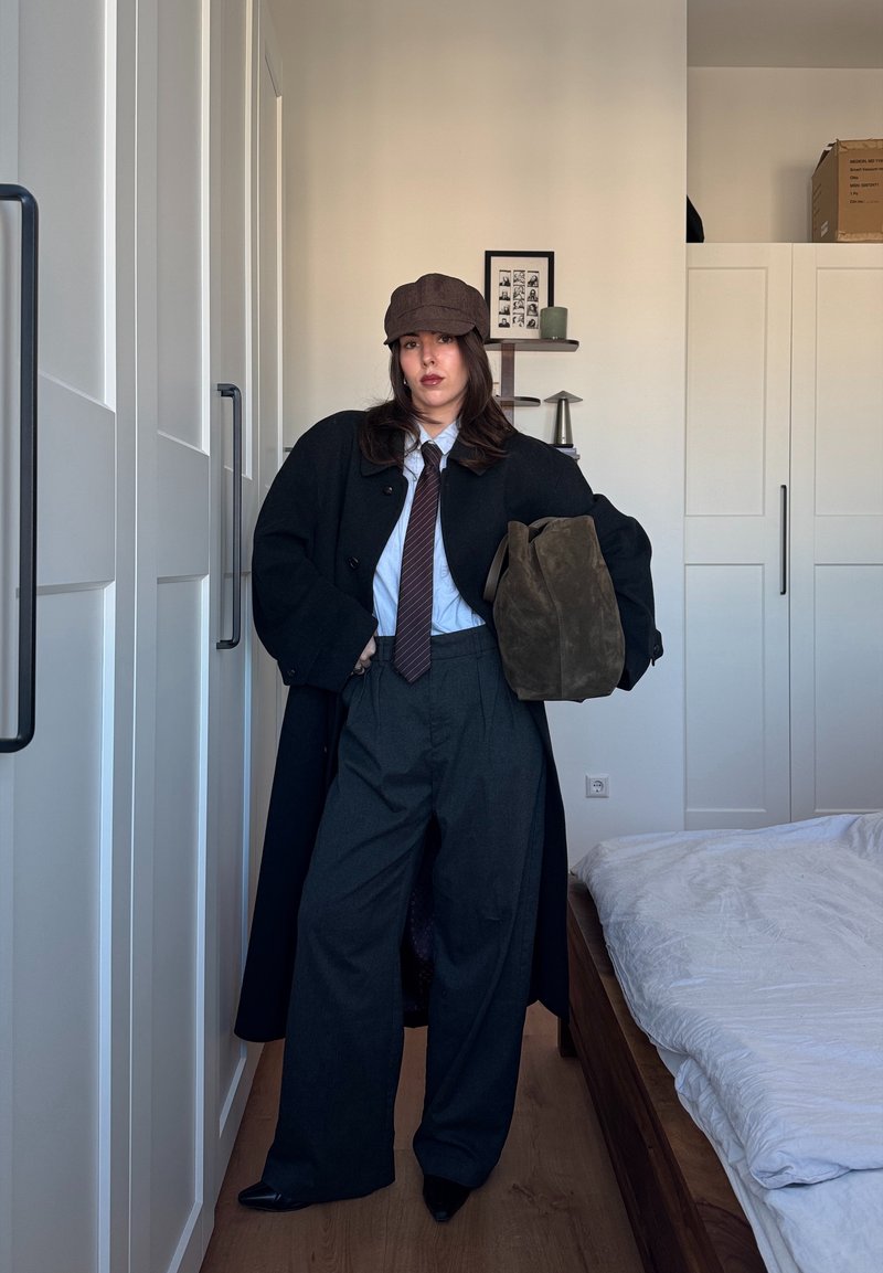 Schwarzer Mantel, hellblaues Hemd, braune Krawatte, übergroße graue Hose, braune Schiebermütze, olivgrüne Tasche, in einer minimalistischen Innenraumszene.