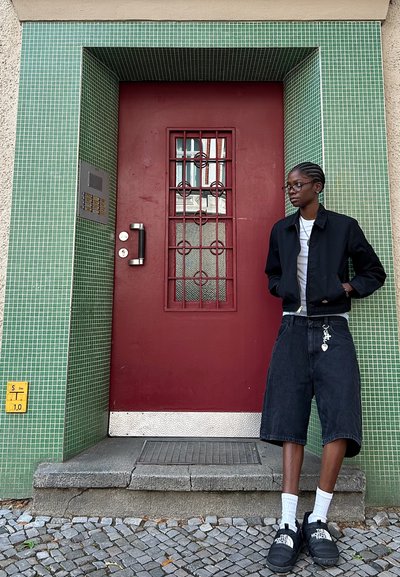 Puerta roja con un pomo de metal y ventana con patrón de rejilla, rodeada de paredes de azulejos verdes. Persona vestida con chaqueta negra y pantalones cortos de mezclilla se encuentra cerca.