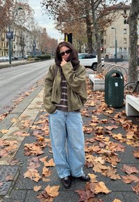 Veste surdimensionnée vert olive, haut rayé marron, jean bleu ample et chaussures foncées. Entouré de feuilles tombées dans une rue de la ville.