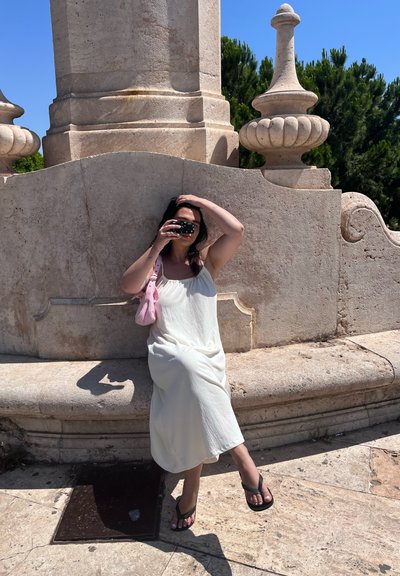 Una persona sentada en un vestido blanco fluido y chanclas negras, sosteniendo una cámara, frente a un fondo de piedra con detalles tallados.