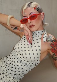 Valge täpilise mustriga kleit, millel on kaelaümber seotud lõige ja kortsuline disain, kombineeritud punaste päikeseprillide ja akrüül-ehetega. Õlal lilleline tätoveering.