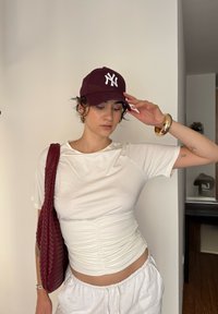 Top blanco ajustado con detalles fruncidos, combinado con pantalones blancos holgados. Una gorra burdeos presenta un logo blanco; está accessorized con un bolso trenzado y joyería.